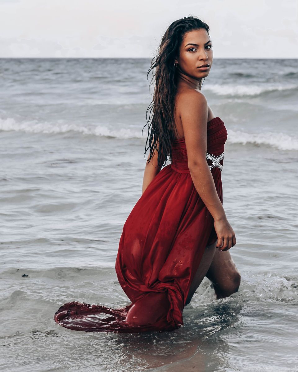 Woman in a flowing maroon off-shoulder maxi dress standing in shallow seawater