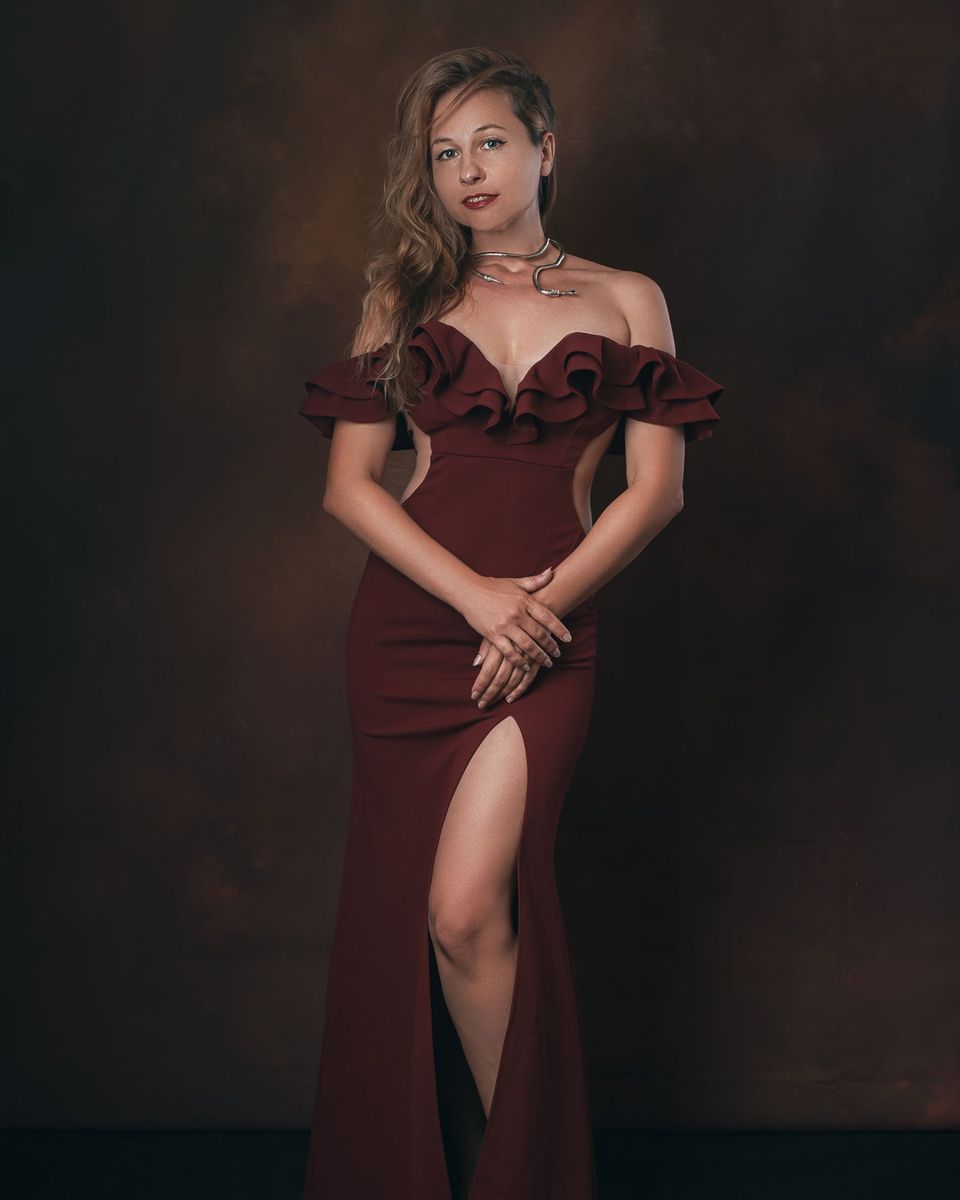 Woman in a maroon off-shoulder evening gown with ruffle details against a dark backdrop